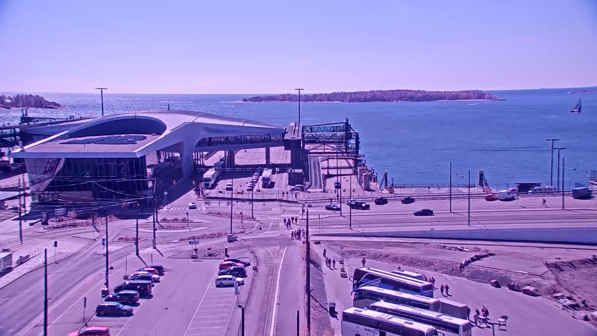 A ferry terminal with vehicles and pedestrians, situated on a waterfront on a clear, sunny day.