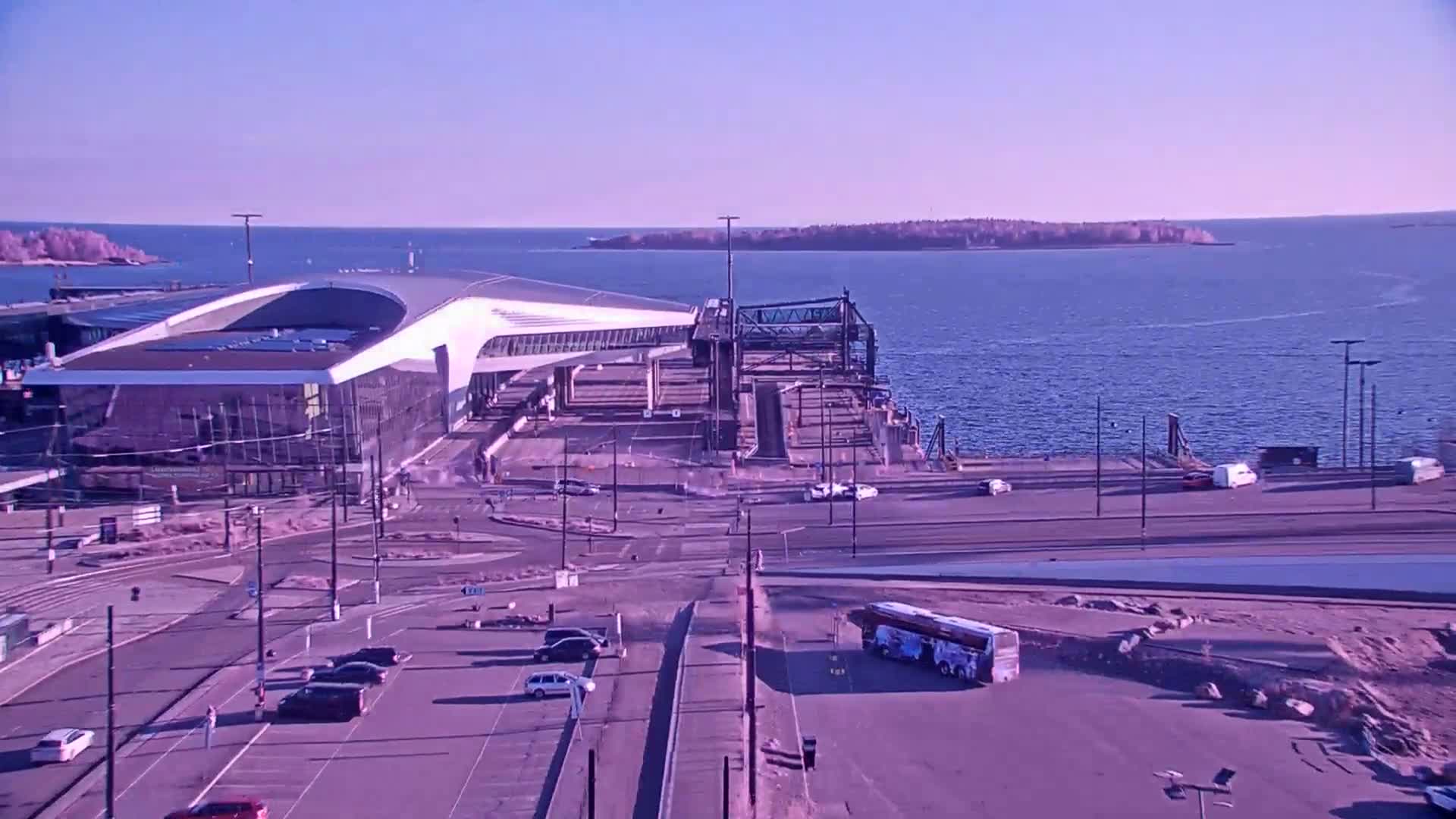 A modern terminal building sits by a calm body of water on a sunny day, with vehicles parked nearby and a bus visible in a lot.