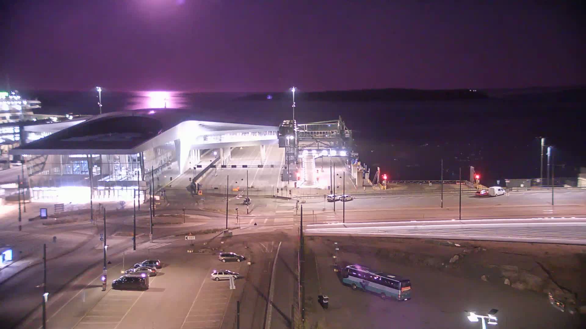 A nighttime view of a ferry terminal with a modern building, cars parked in lots, and a bus, under a purple-tinged sky.