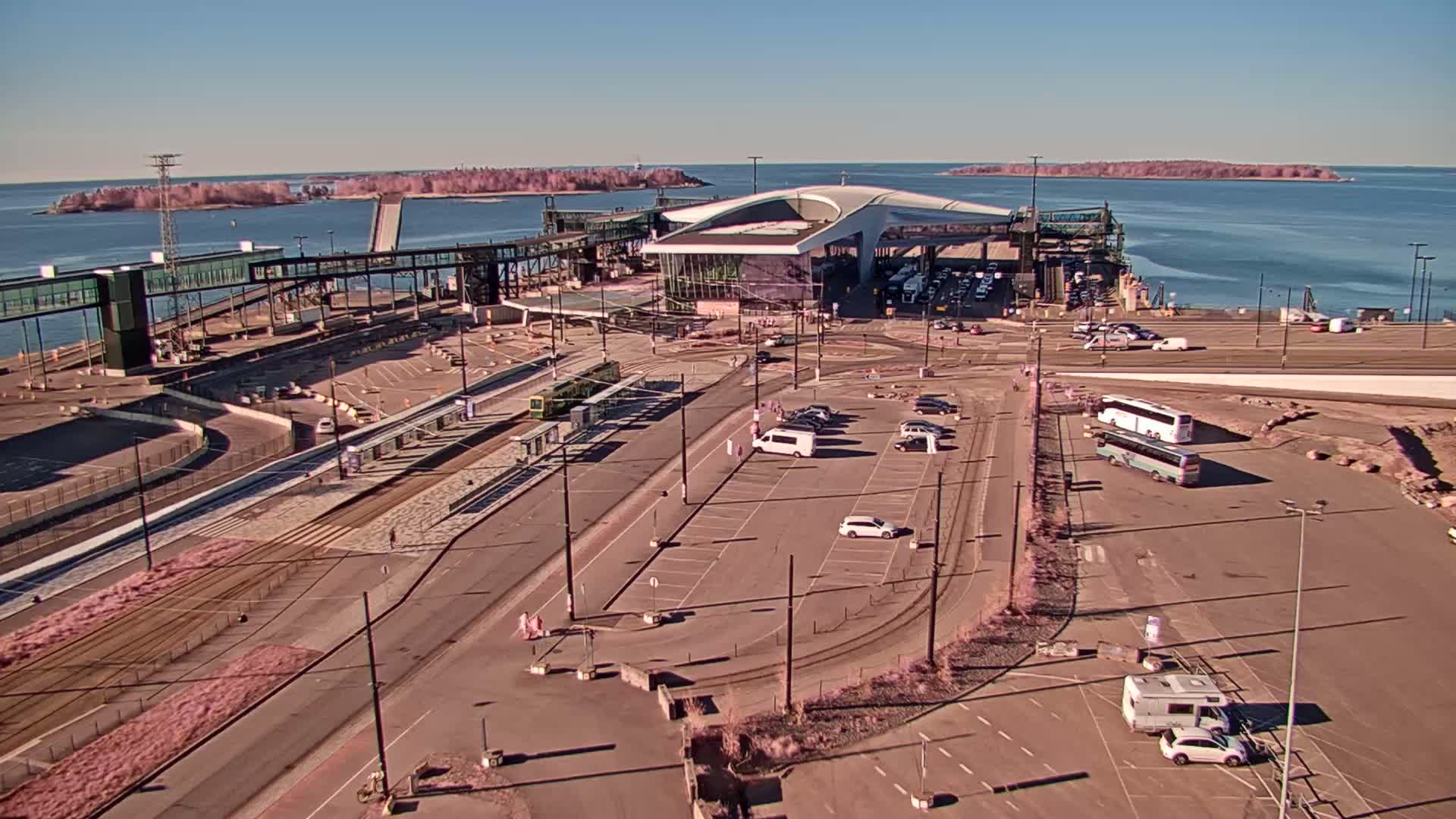 A ferry terminal with a modern building, trams, buses, and cars parked in a large lot, on a sunny day with calm water and distant islands visible.