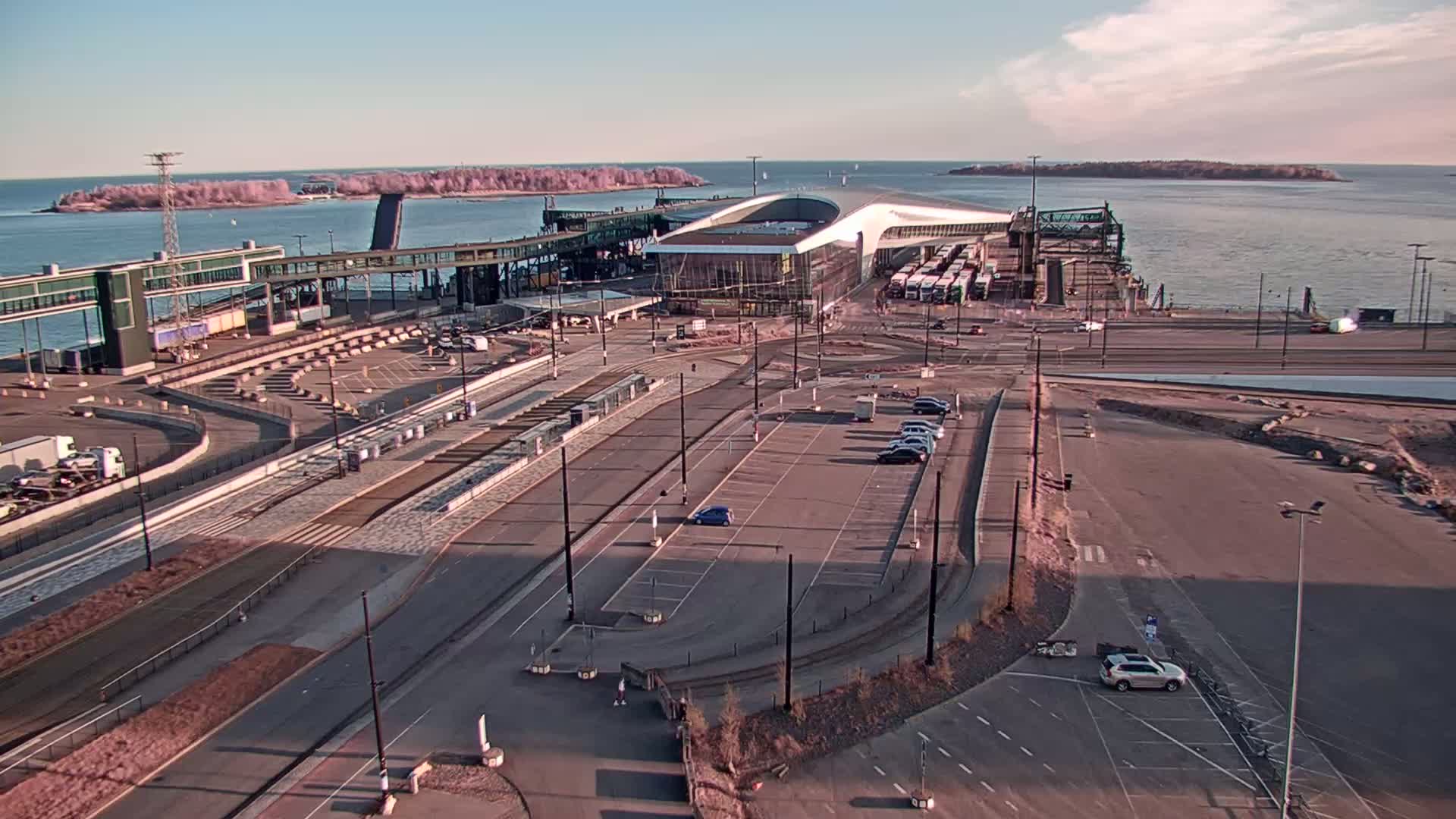 A ferry terminal with a large modern building, numerous vehicles, and an empty parking lot is seen on a sunny day next to calm water with small islands in the distance.