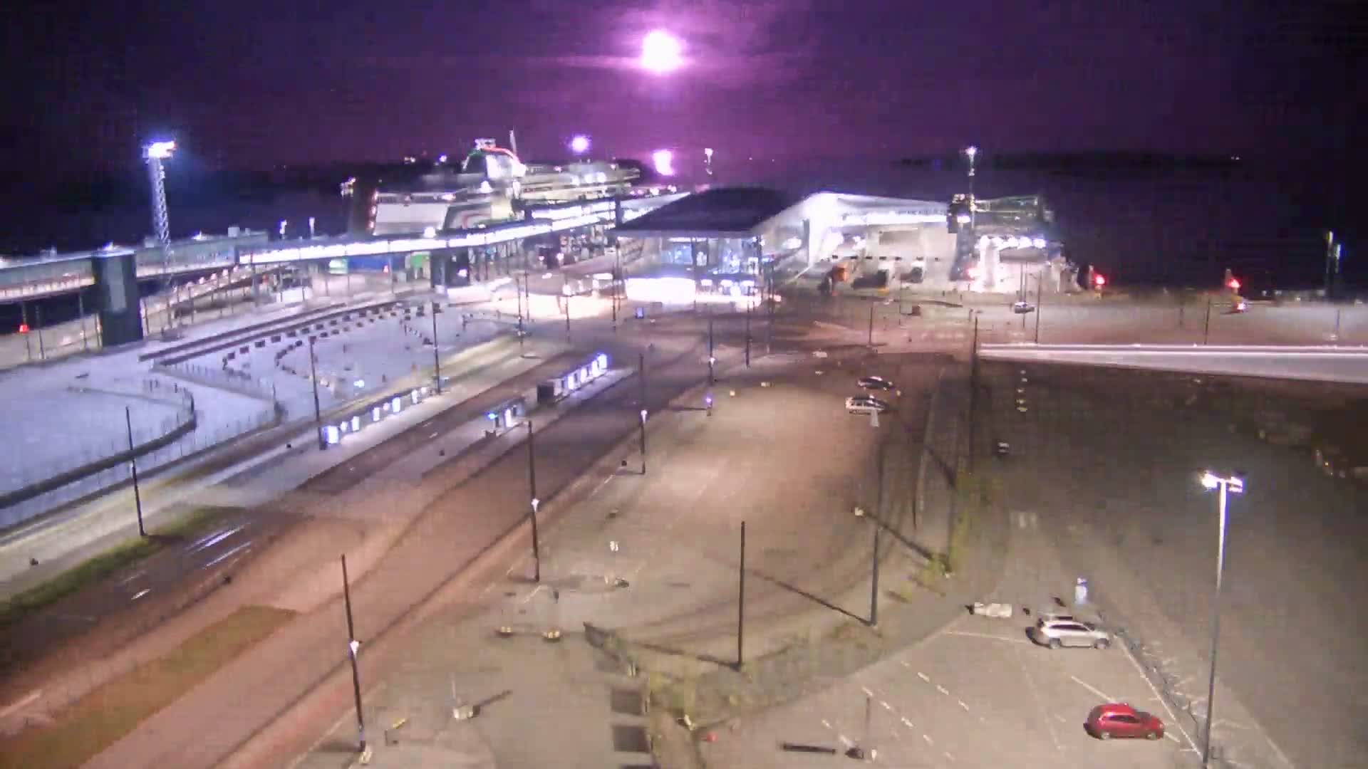 A cruise ship is docked at a terminal under a twilight sky, with several cars parked in a lot nearby, and tram tracks running through the scene.