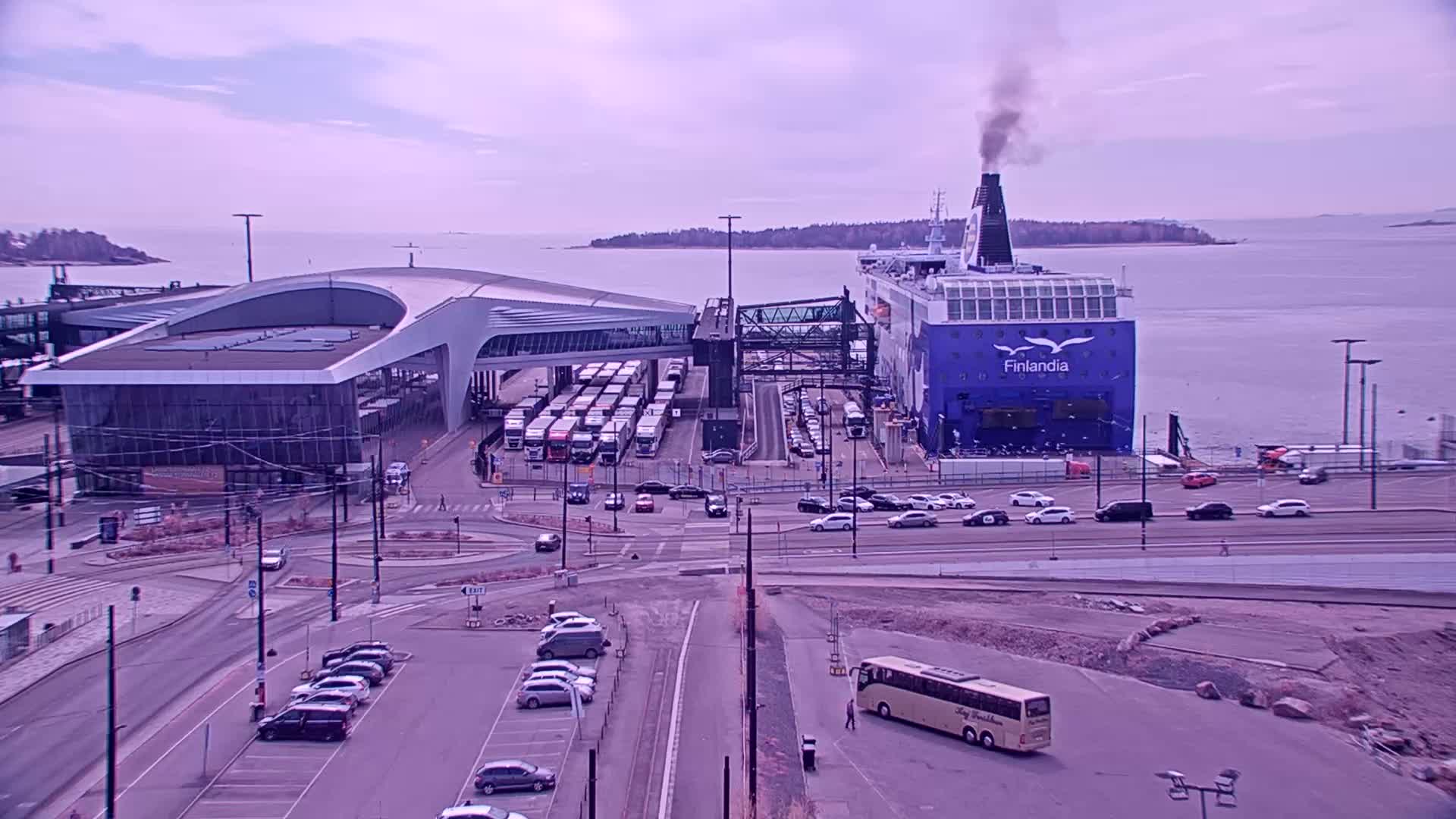 A large ferry is docked at a terminal building, with numerous cars and buses in the surrounding parking lots and roadways under a cloudy sky.