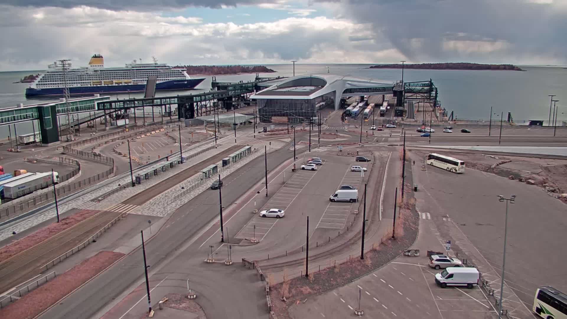 A cruise ship is docked at a terminal, with several cars and buses parked nearby under partly cloudy skies.