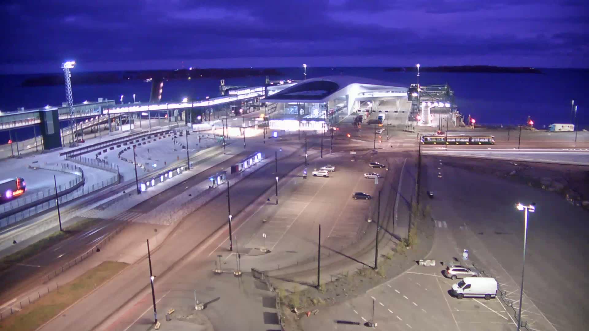 A ferry terminal at night, with a few cars and a tram in the parking lot and surrounding areas, under a partly cloudy, dark blue sky.