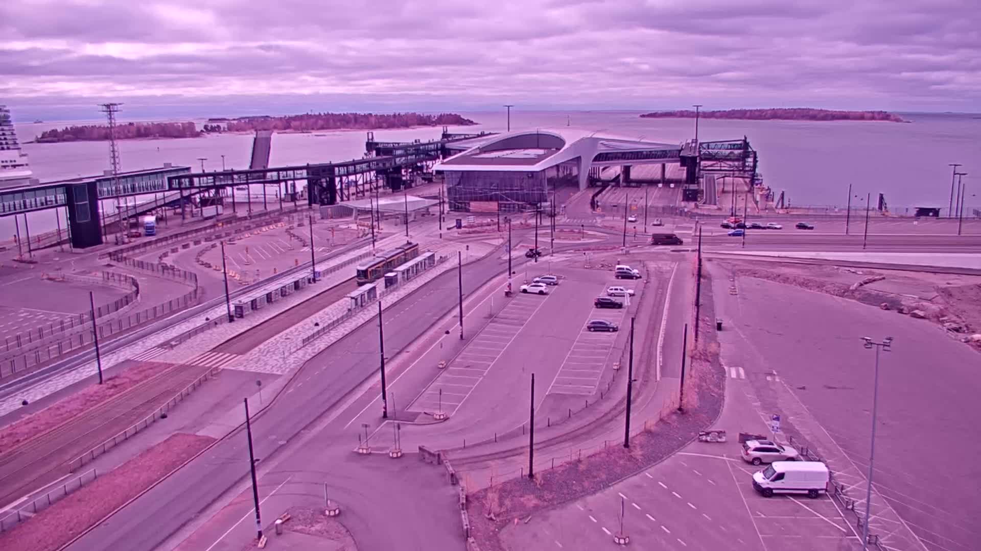 An aerial view of a ferry terminal with a tram, cars in a parking lot, and a calm body of water under a cloudy, purplish sky.