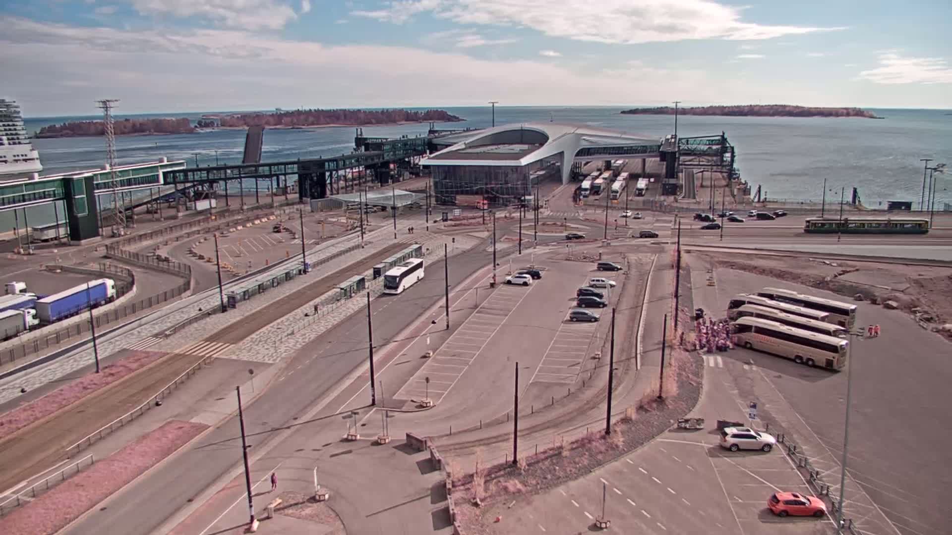 An aerial view of a ferry terminal on a sunny day, showing buses, cars, and a passenger ferry docked at the terminal.