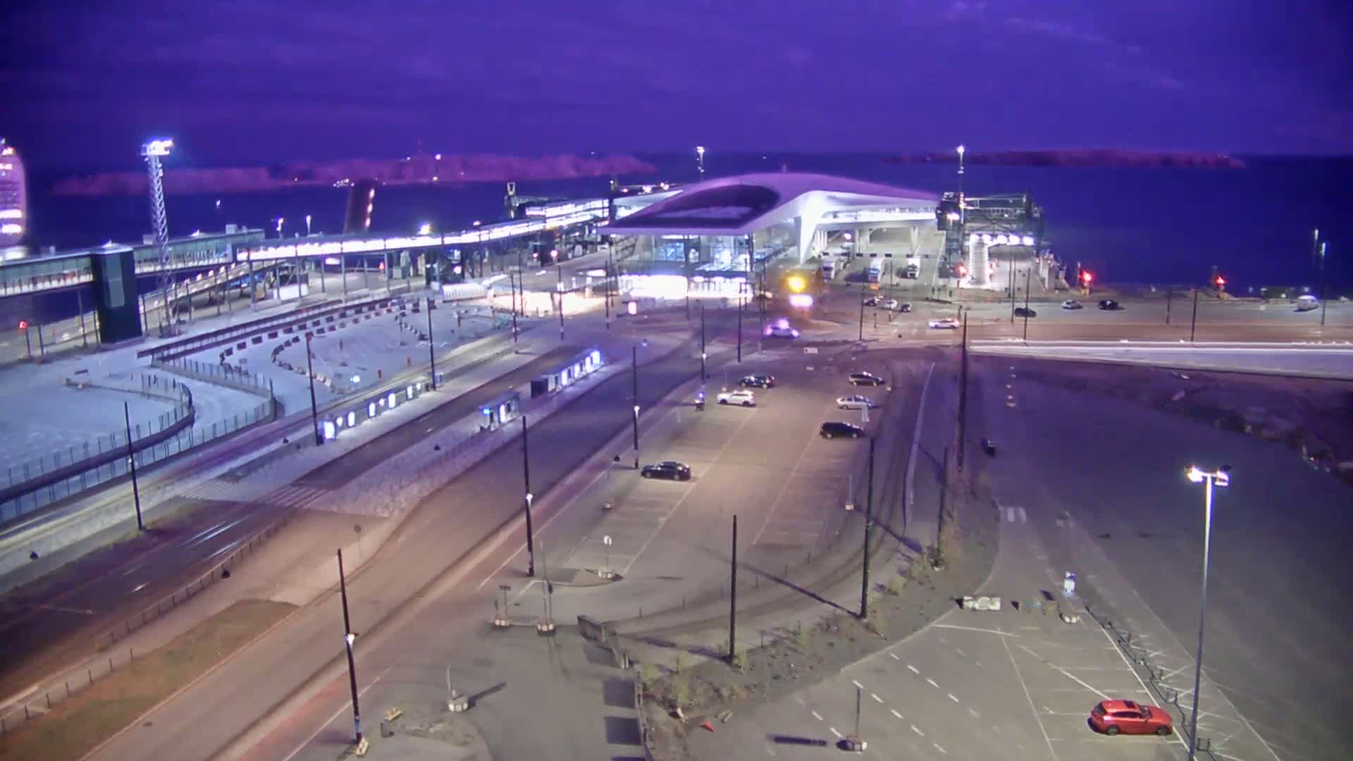A ferry terminal at night under a twilight sky, with several cars parked in a lot nearby.