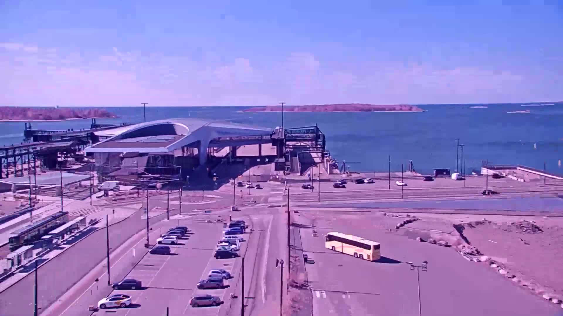 A modern ferry terminal sits by a calm body of water on a sunny day, with several vehicles parked nearby and a bus in a parking lot.
