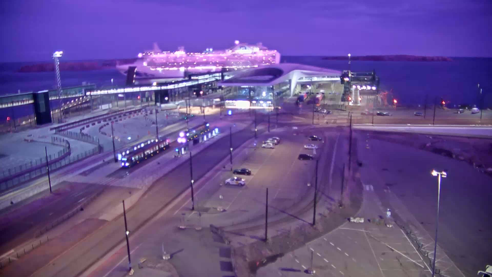 A large cruise ship is docked at a terminal, with a tram and cars visible in a parking lot under a twilight sky.