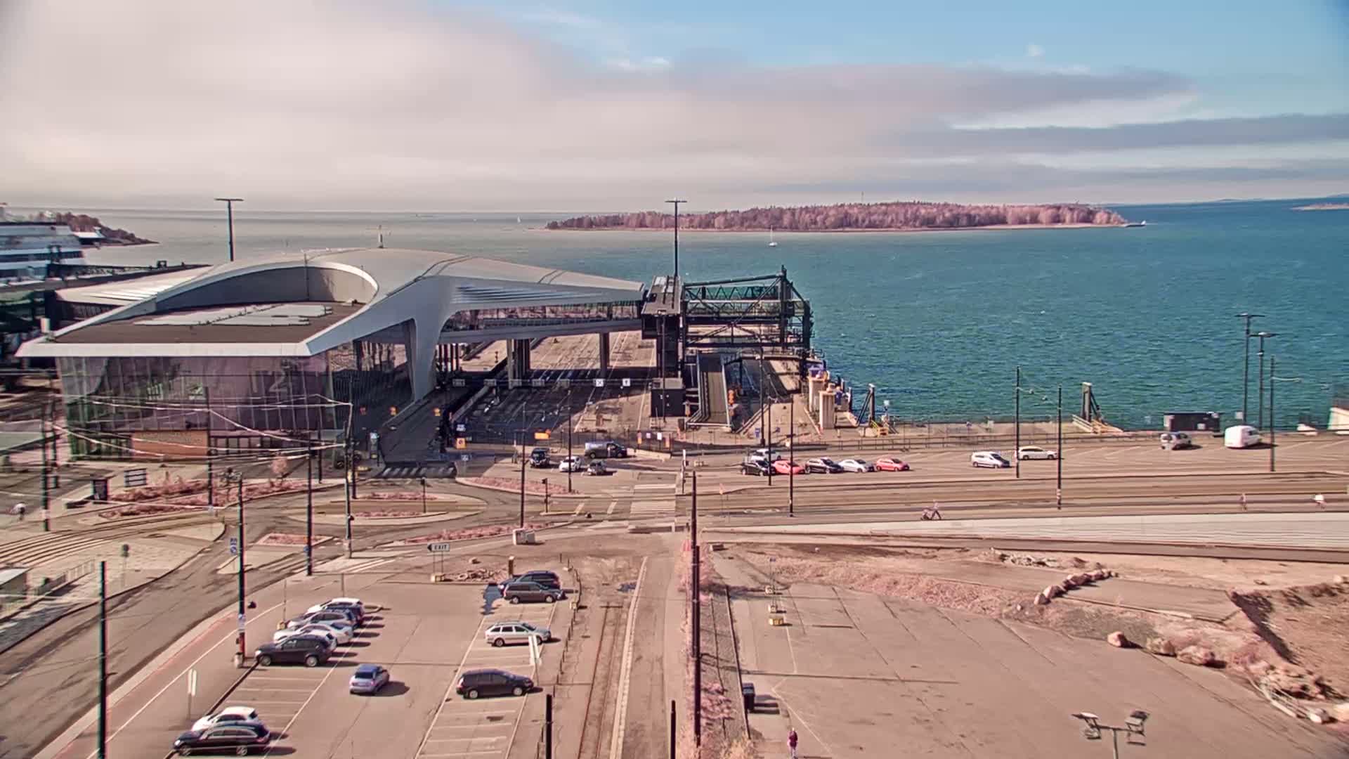A modern ferry terminal building sits on a waterfront, with cars parked nearby under a partly cloudy sky.