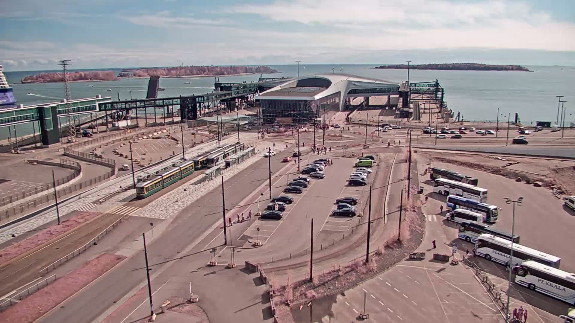 A ferry terminal with a tram, cars, and buses parked, on a sunny day with a calm body of water and islands in the background.