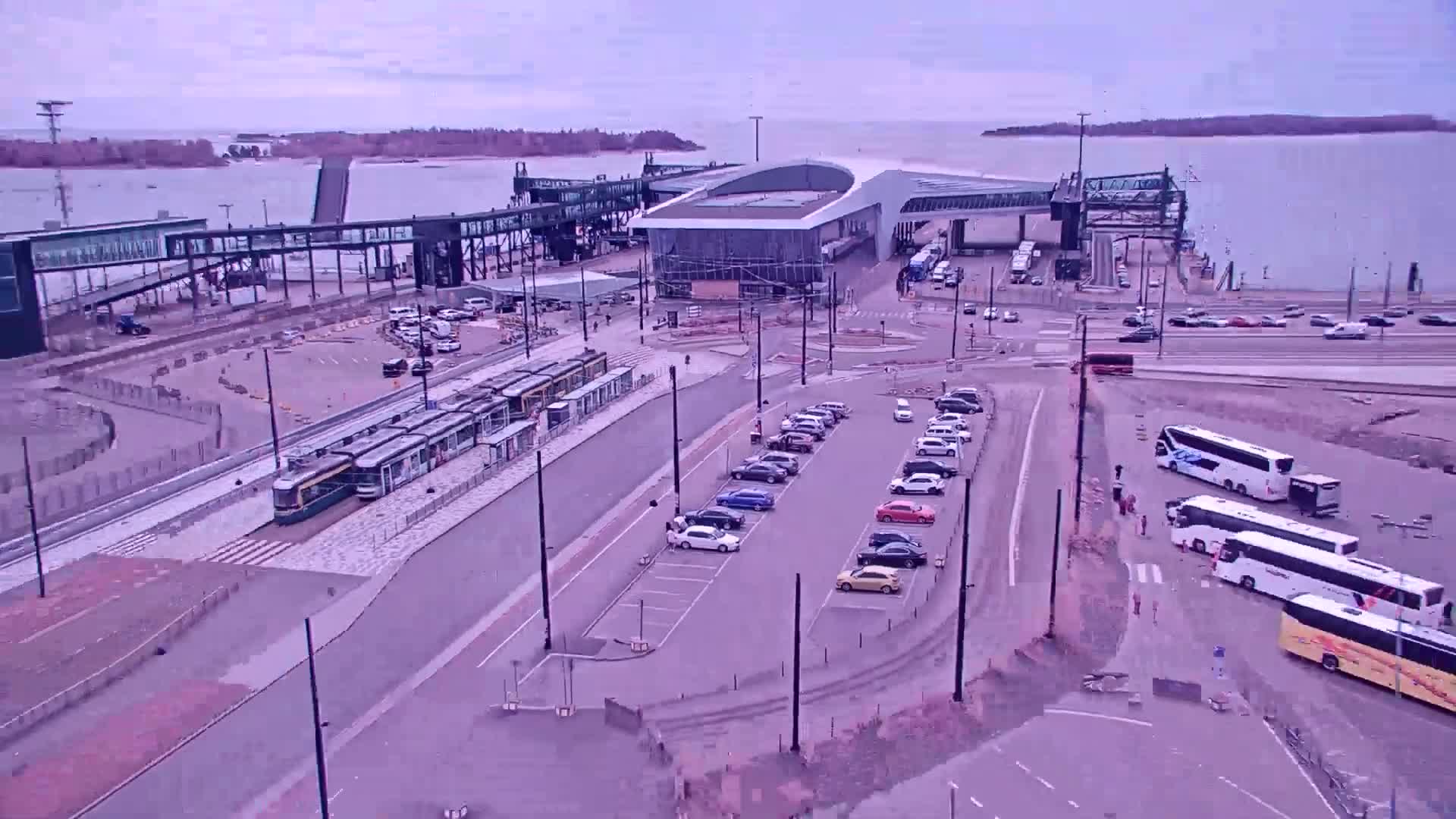 An aerial view of a ferry terminal on an overcast day shows trams, cars parked in a lot, and several tour buses near the terminal building.
