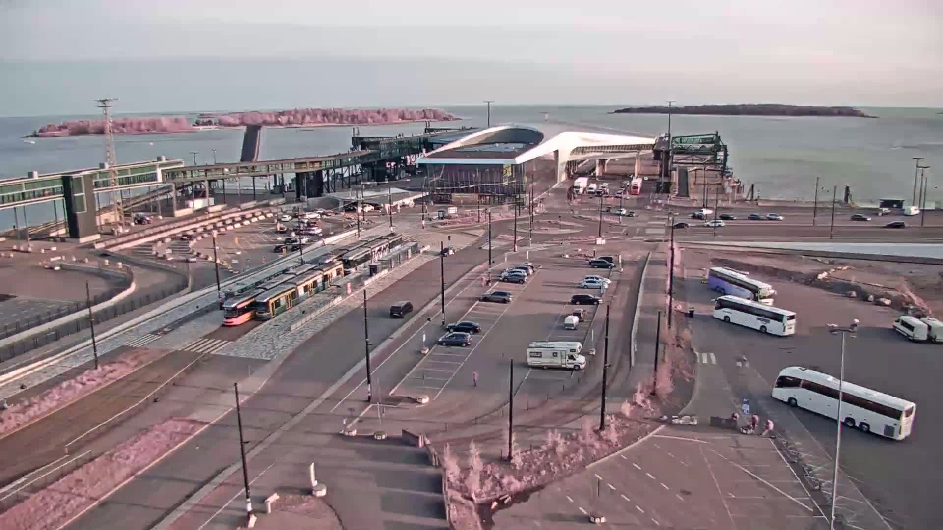 An aerial view shows a ferry terminal with a tram, several cars and buses in a parking lot, and calm water with islands in the background under a clear sky.