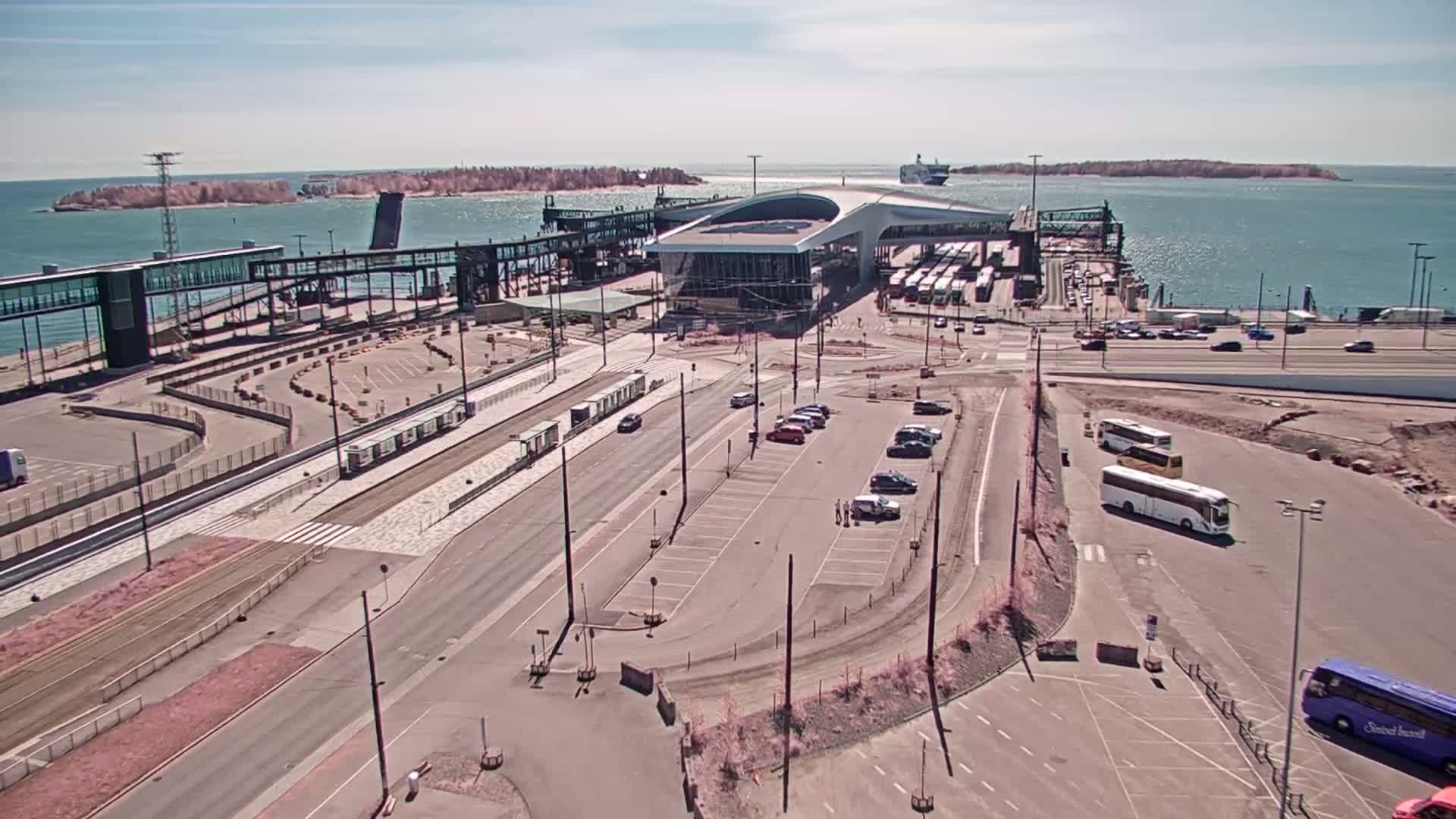 An aerial view of a ferry terminal on a sunny day, showing a large terminal building, numerous vehicles, buses, and a ferry in the distance.