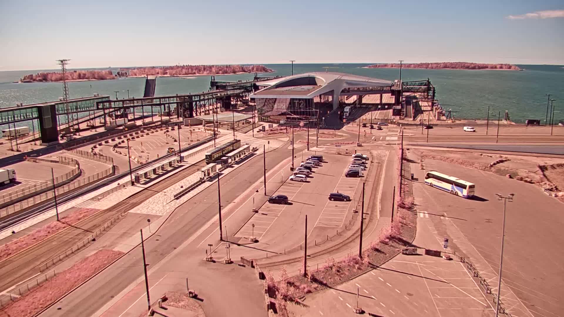 An aerial view shows a ferry terminal on a sunny day, with trams, cars, and a bus parked nearby, and islands visible in the distance across the water.