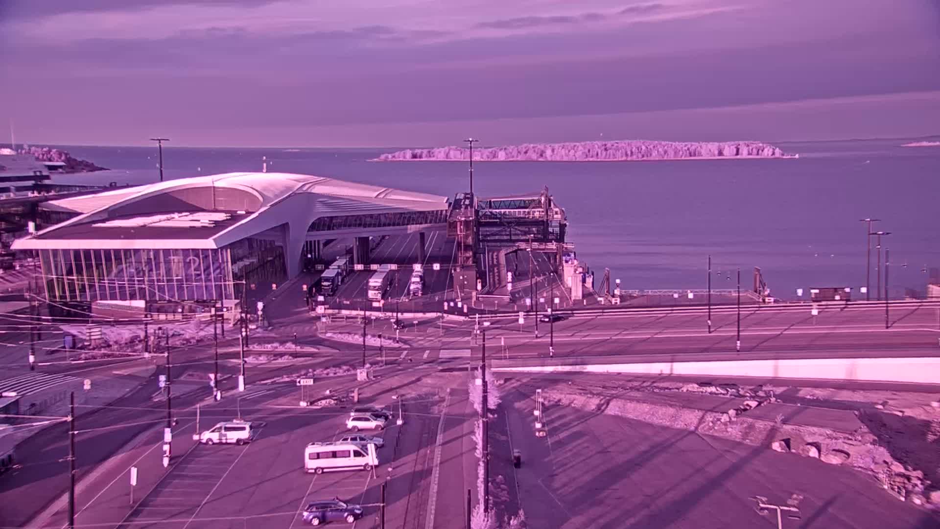 A modern ferry terminal with several vehicles parked nearby overlooks a calm body of water and a distant island under a purplish twilight sky.