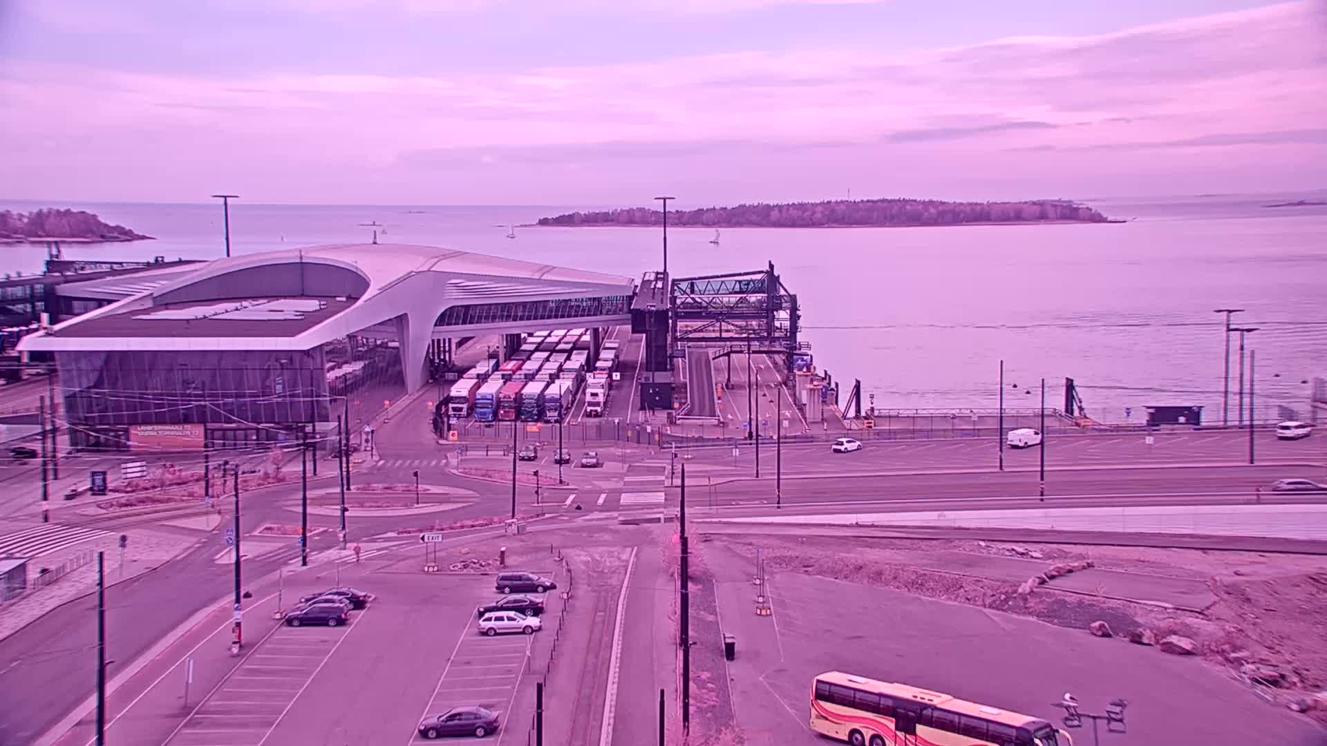 A ferry terminal with numerous semi-trailer trucks and a few cars is shown under a pale purple sky, next to a body of water.