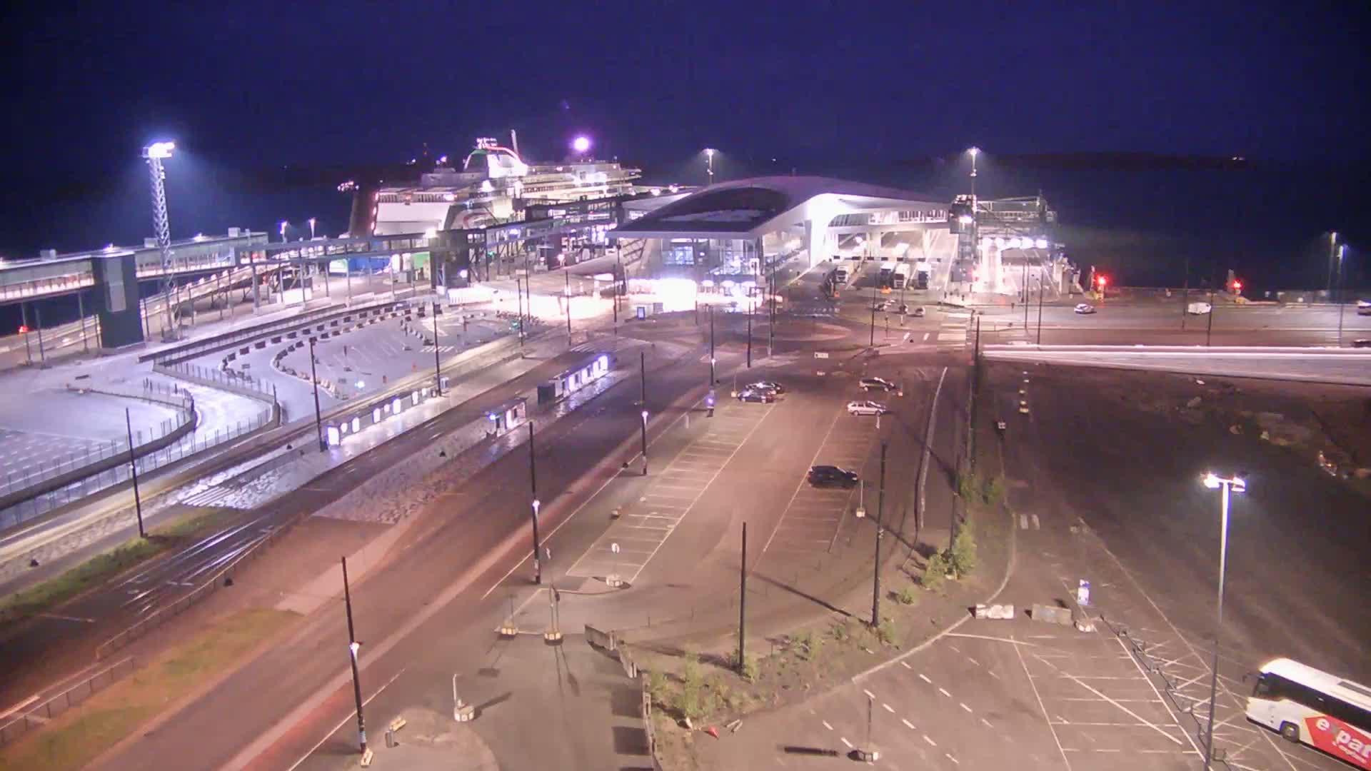 A ferry terminal at night, illuminated by artificial lights, with a large ferry docked and several vehicles in the parking lot.