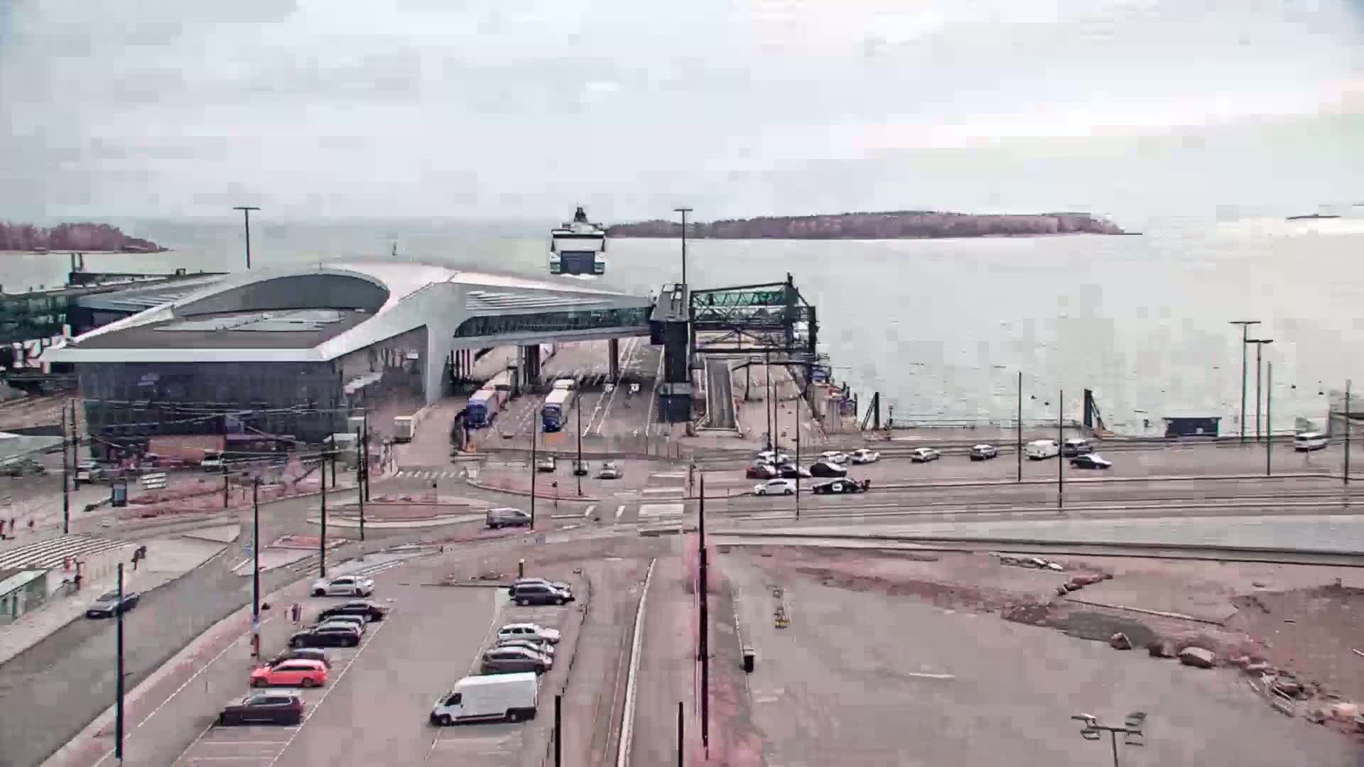 A ferry terminal with a large building and connecting walkway, cars in a parking lot, and a ferry docked, under an overcast sky.