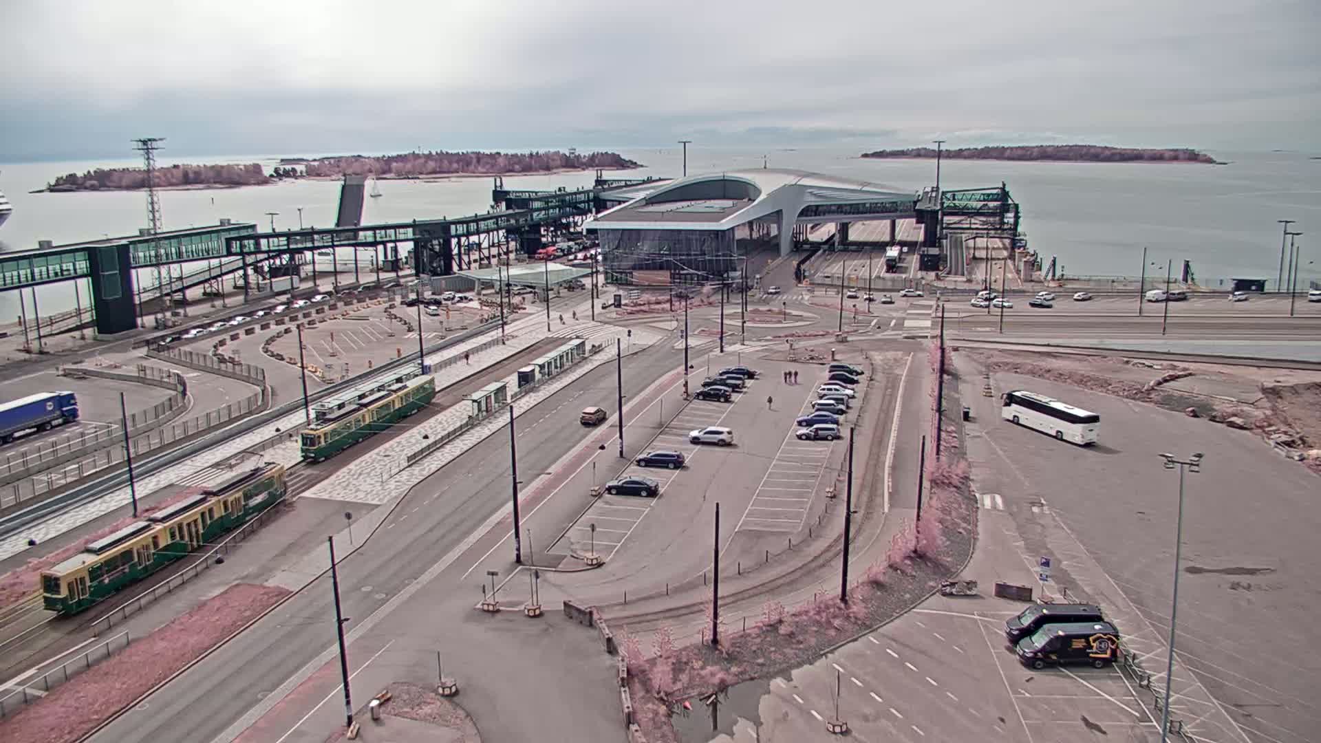 An aerial view shows a ferry terminal with two trams, several cars in a parking lot, a bus, and a partly cloudy sky.