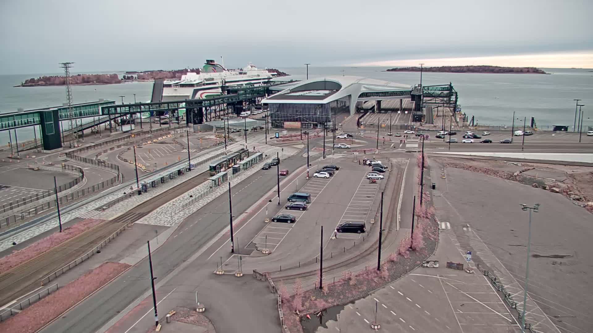 An overcast day at a ferry terminal shows a large ferry docked, connected to a modern terminal building by a walkway, with trams and cars in the surrounding parking lots and roadways.
