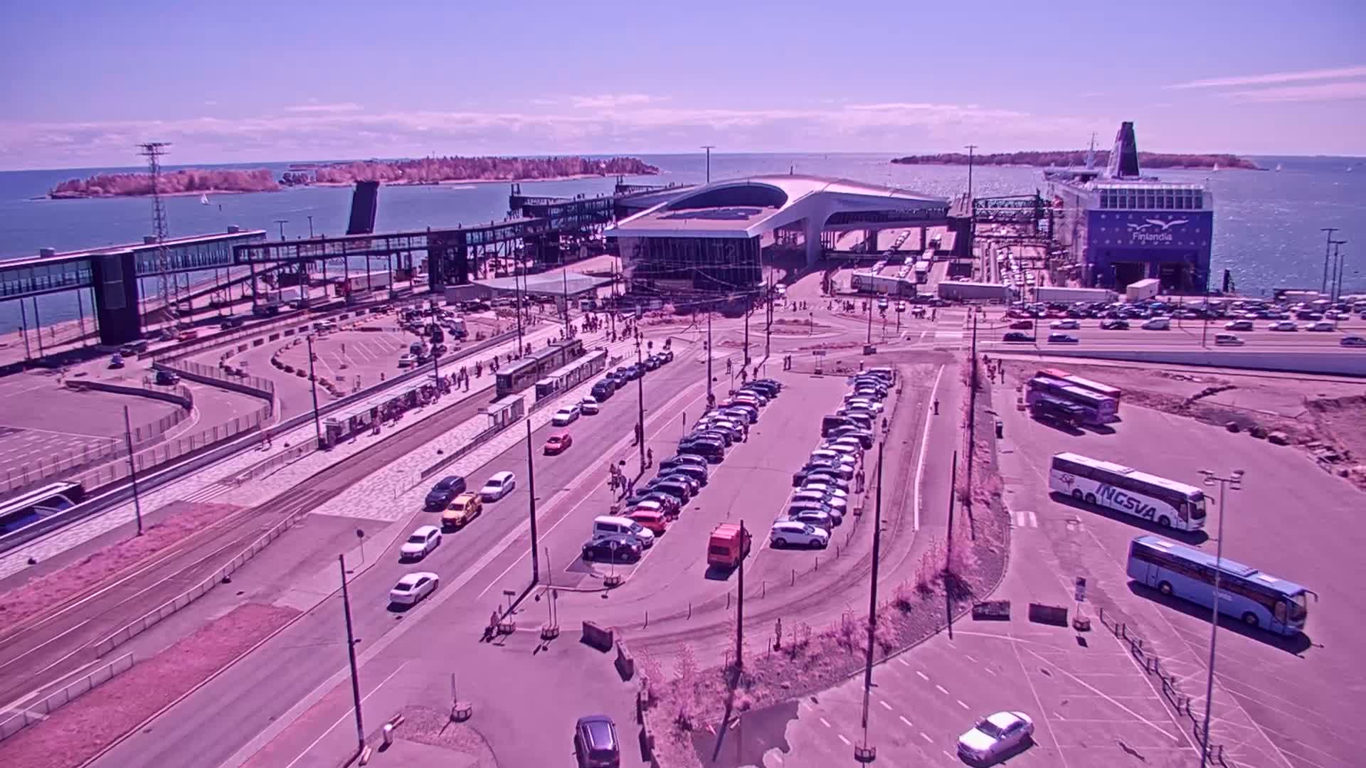 A ferry is docked at a terminal on a sunny day, with cars and buses parked nearby and pedestrians walking around.