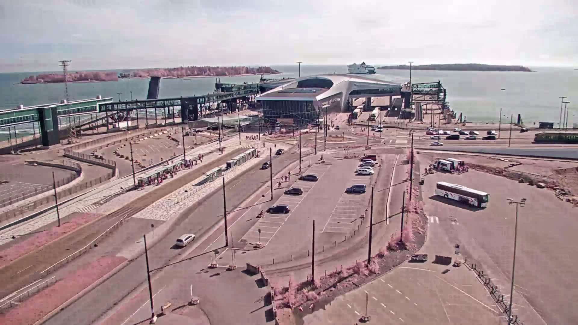 An aerial view of a ferry terminal on a sunny day shows a large parking lot, a bus, several cars, and pedestrians near a ferry boat docked at the terminal.