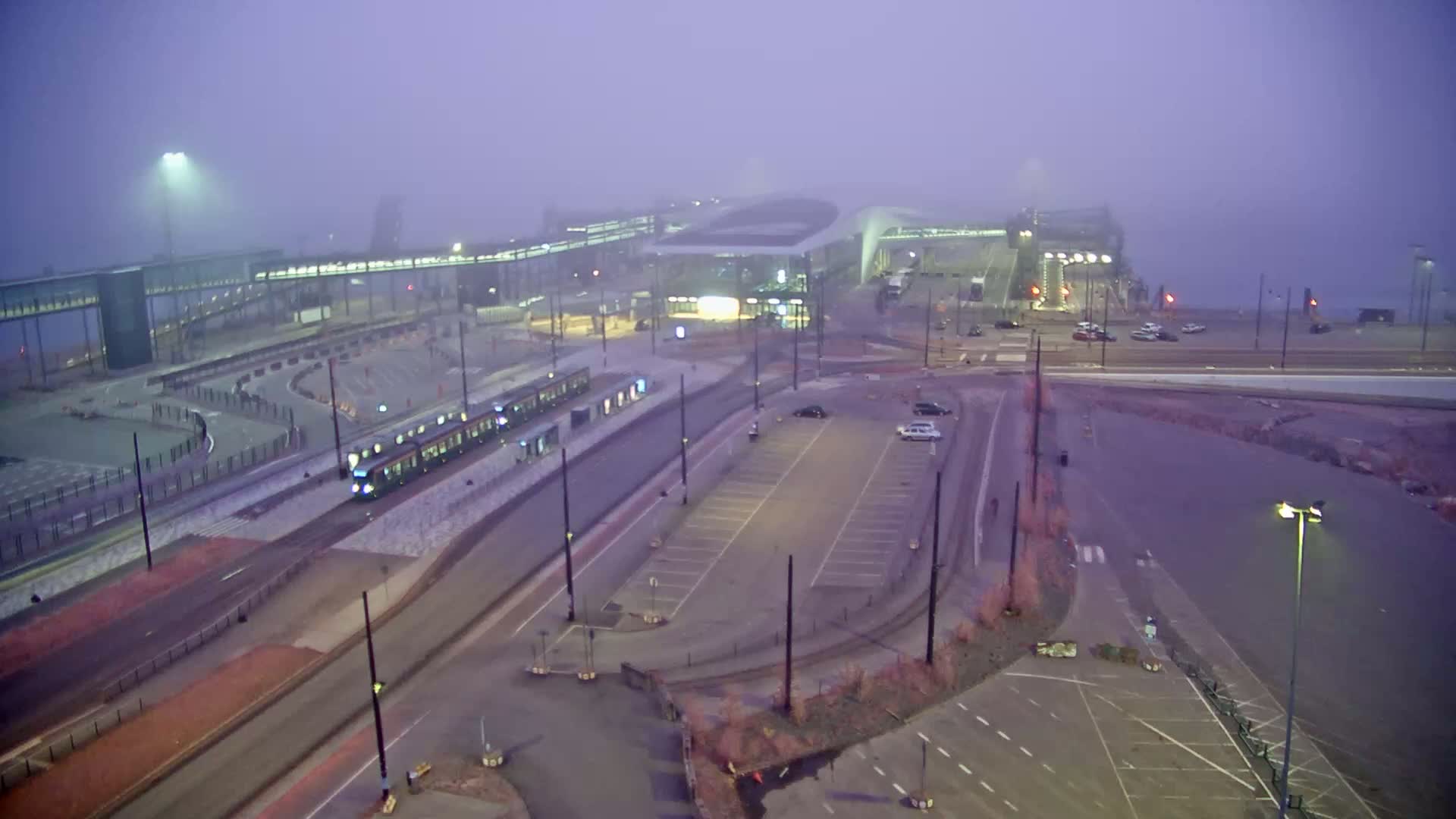 A tram is at a station next to a large parking lot and a ferry terminal, all shrouded in thick fog.