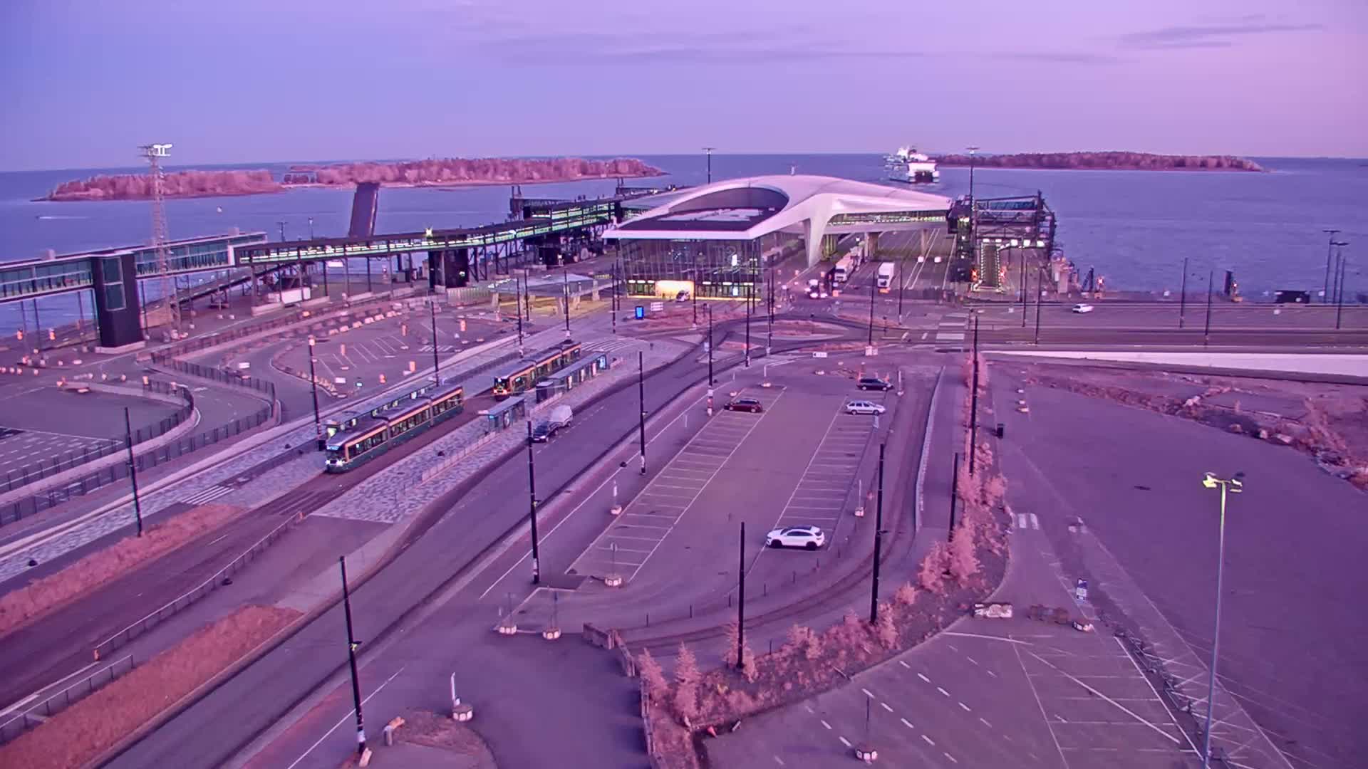 An aerial view of a ferry terminal at dusk, with two trams at a station, a few cars in a parking lot, and a ferry in the water.