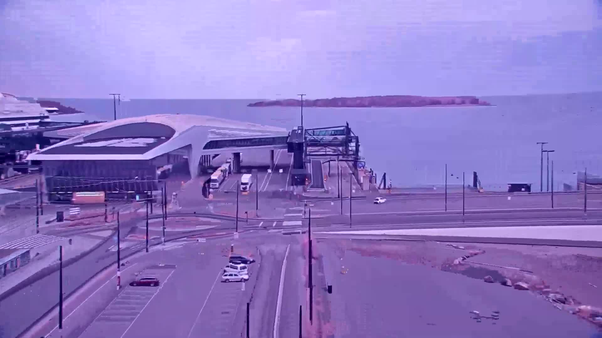 A ferry terminal with a modern building and a loading bridge sits on a body of water under an overcast sky.