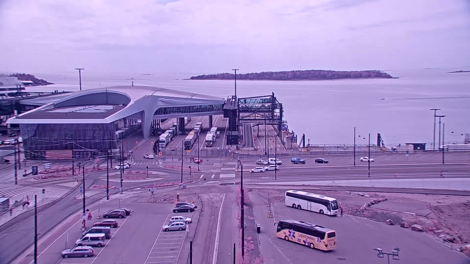 A ferry terminal with several buses and cars in a parking lot, under an overcast sky, next to a body of water with an island in the distance.