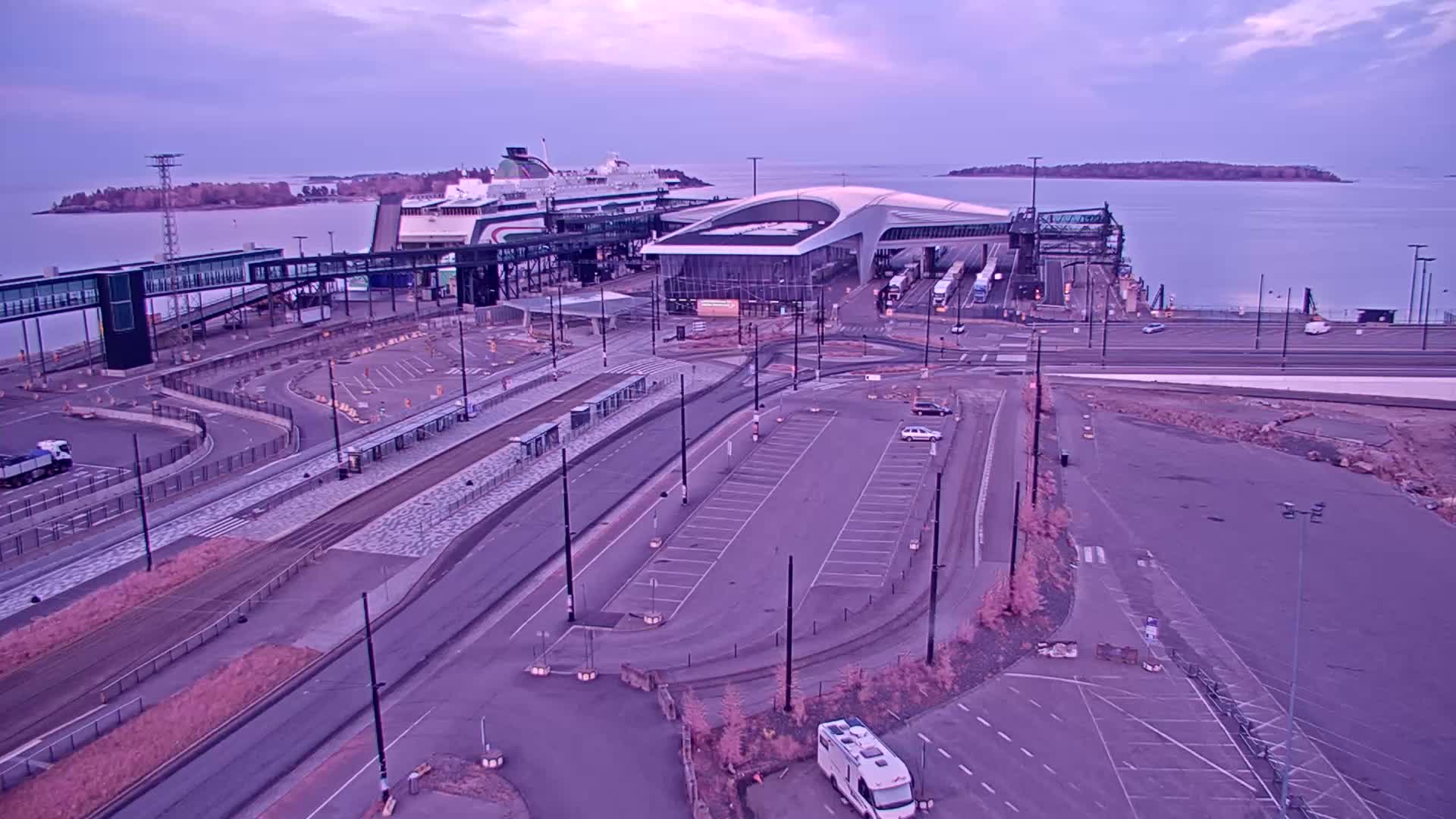 An aerial view of a ferry terminal at dusk, with a large ferry docked and a nearly empty parking lot.