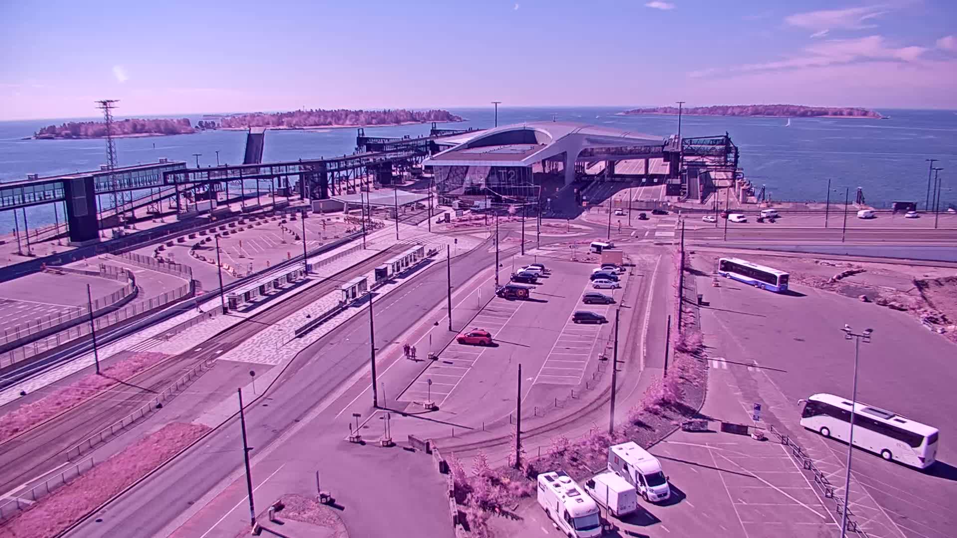 An aerial view of a ferry terminal on a sunny day, showing a modern terminal building, parking lots, and several buses and cars.