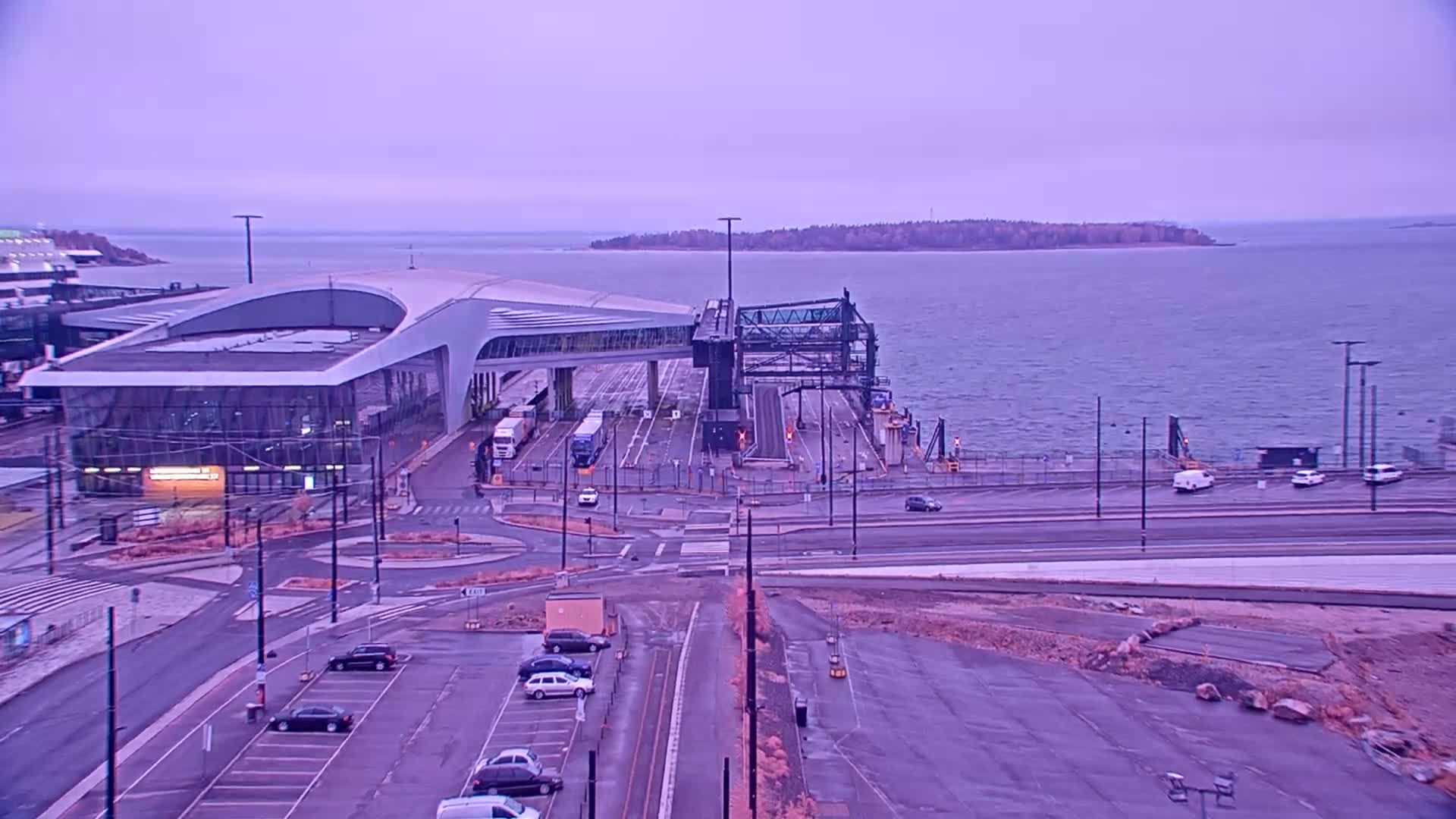 A ferry terminal with a modern building and loading docks sits by a calm body of water under a pale purple sky.