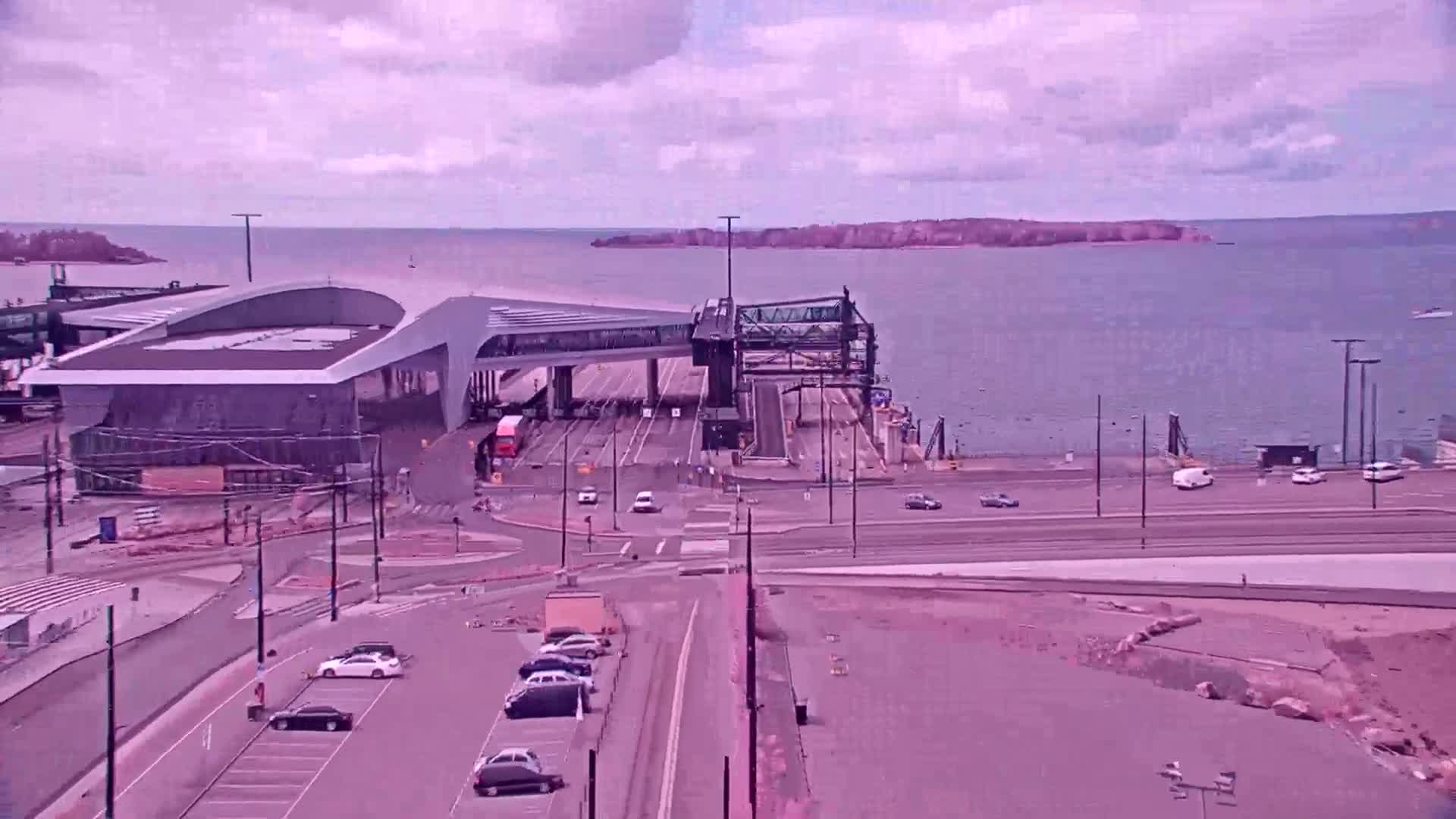 A ferry terminal with a modern building and a connecting walkway overlooks a calm body of water under a mostly cloudy sky.