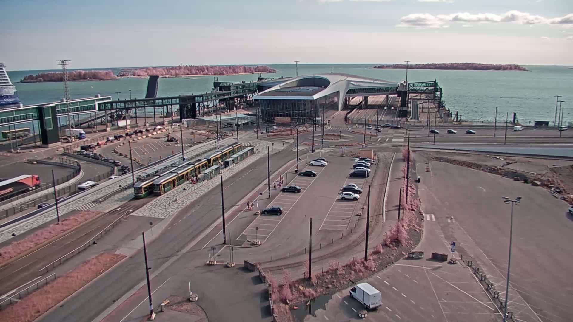 A ferry terminal with trams and parked cars is seen on a sunny day next to calm water and distant islands.