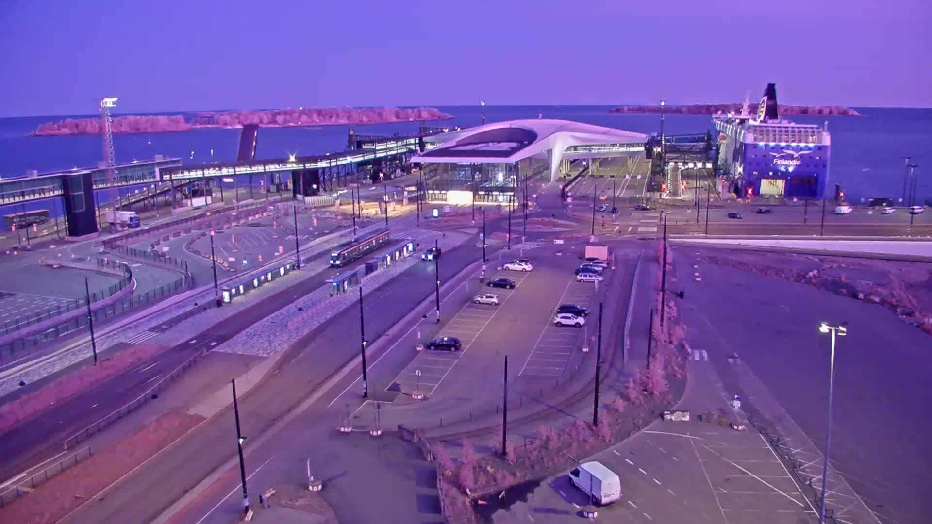 A ferry is docked at a terminal with a modern terminal building, parking lots, and roadways under a twilight sky.