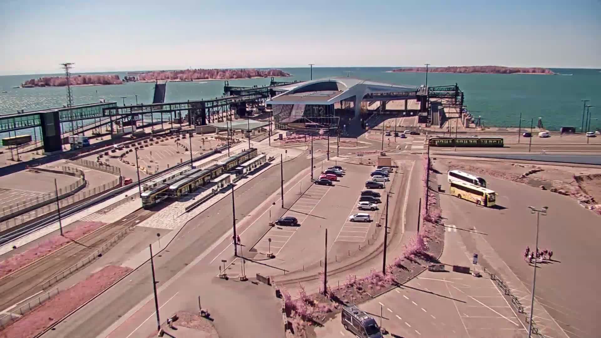 A sunny day overlooks a ferry terminal with trams, buses, and cars parked nearby, across from a body of water with several small islands in the distance.