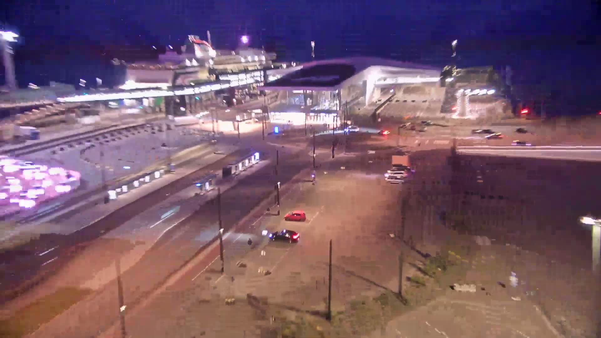 A nighttime aerial view shows a city intersection with several cars, a large stadium, and a modern building, all under dark, clear skies.