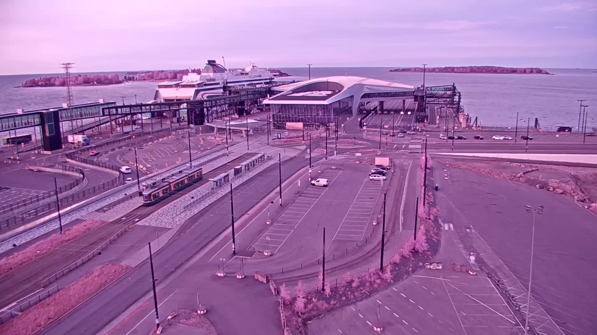 An aerial view shows a ferry terminal with a docked ferry, a tram on tracks, a parking lot, and calm water under a pale sky.