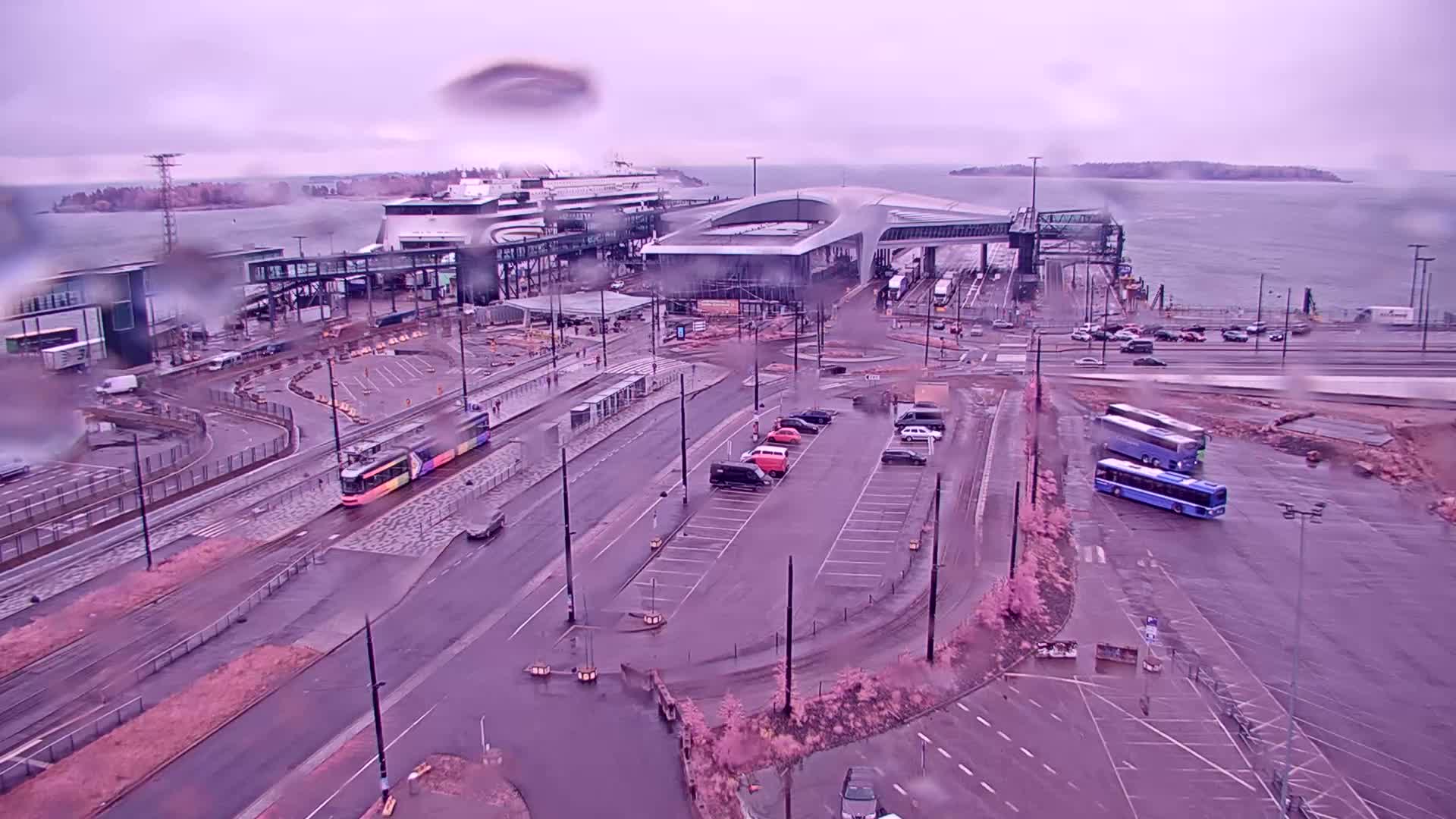 A rainy view of a ferry terminal with a tram, buses, and cars parked in a lot, overlooking a body of water and distant land.
