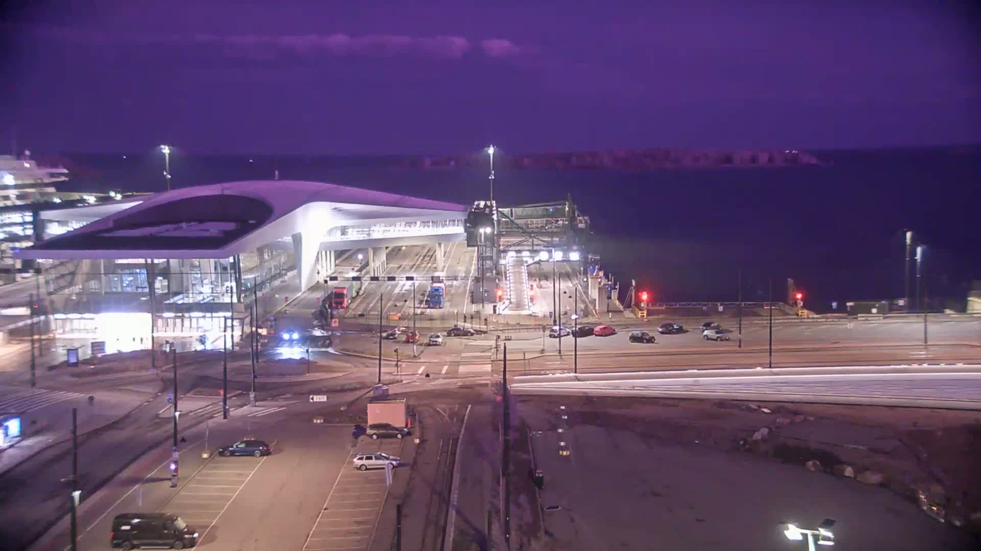 A ferry terminal at night under a purple twilight sky, with several cars parked nearby and a few driving on the roads.