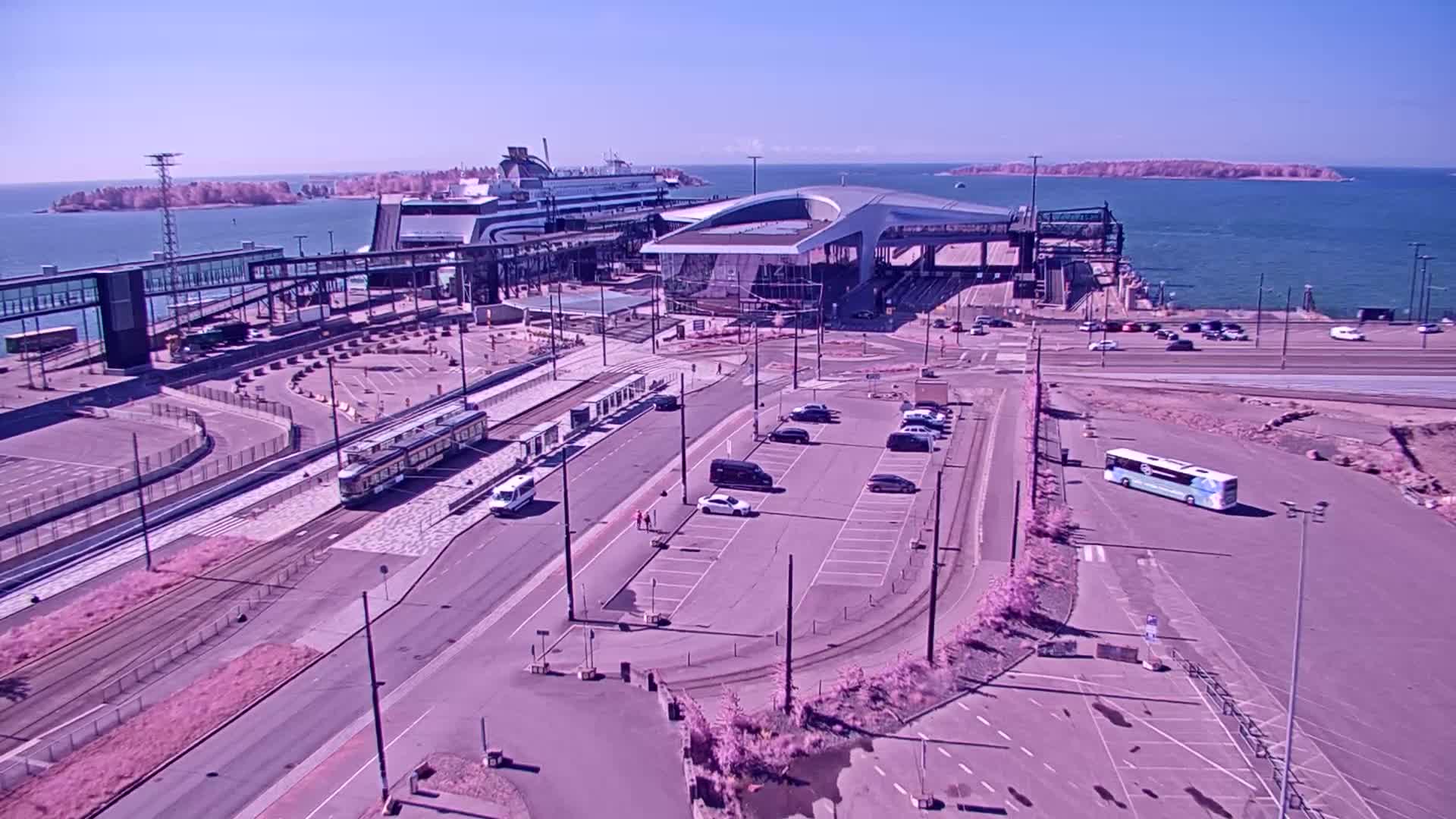 A ferry terminal with a docked ferry, a tram, buses, and cars parked in a large lot on a sunny day.