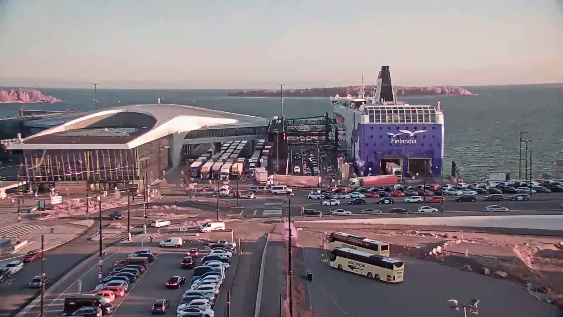 A large ferry named Finlandia is docked at a terminal, surrounded by numerous cars and trucks, under a mostly clear sky.