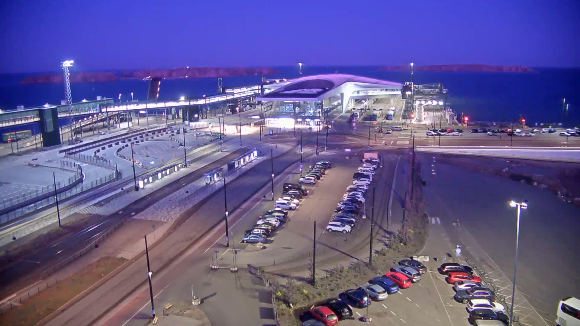 A ferry terminal at night under a dark purple twilight sky, with several cars parked in lots nearby.