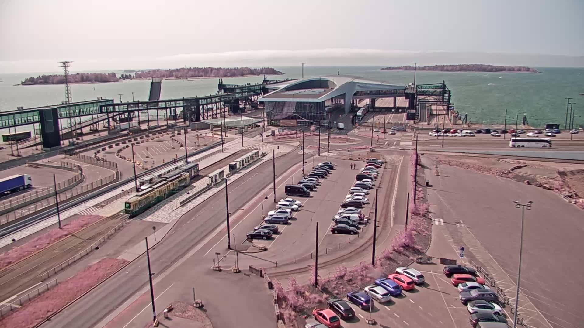 An aerial view shows a ferry terminal with a tram, cars parked in lots, and a calm body of water with islands in the distance under a bright, sunny sky.