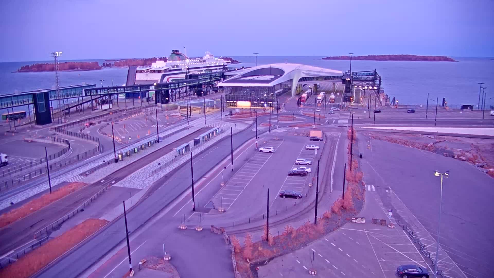 An aerial view of a ferry terminal at dusk, showing a large ferry docked, numerous vehicles in parking lots, and a calm body of water with islands in the background.