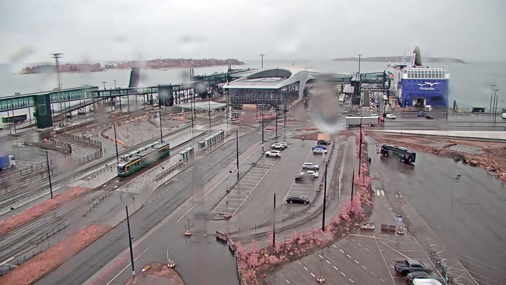 A ferry is docked at a port in rainy weather, with a tram and several cars visible in the surrounding area.