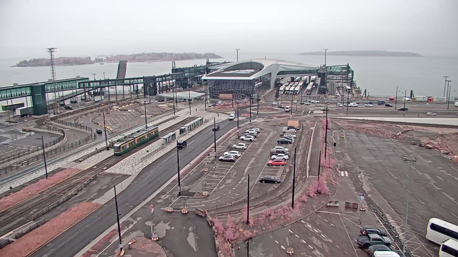 A ferry terminal with a tram, cars in a parking lot, and buses near a body of water on an overcast day.